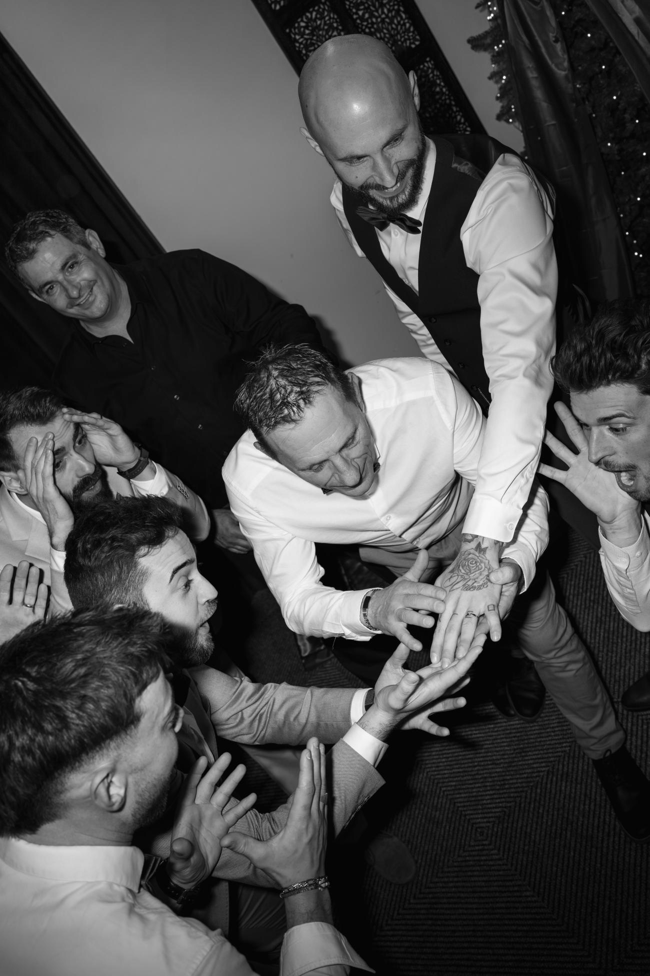 Photo spontanée et drôle du marié et de ses garçons d'honneur. La photo est en noir et blanc de type éditoriale, en contre plongée et ils regardent avec étonnement et éclats de rire l'alliance du marié. 